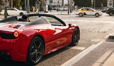 person sitting on the driver's seat of a red convertible coupe at the side of the road near cars on road during day